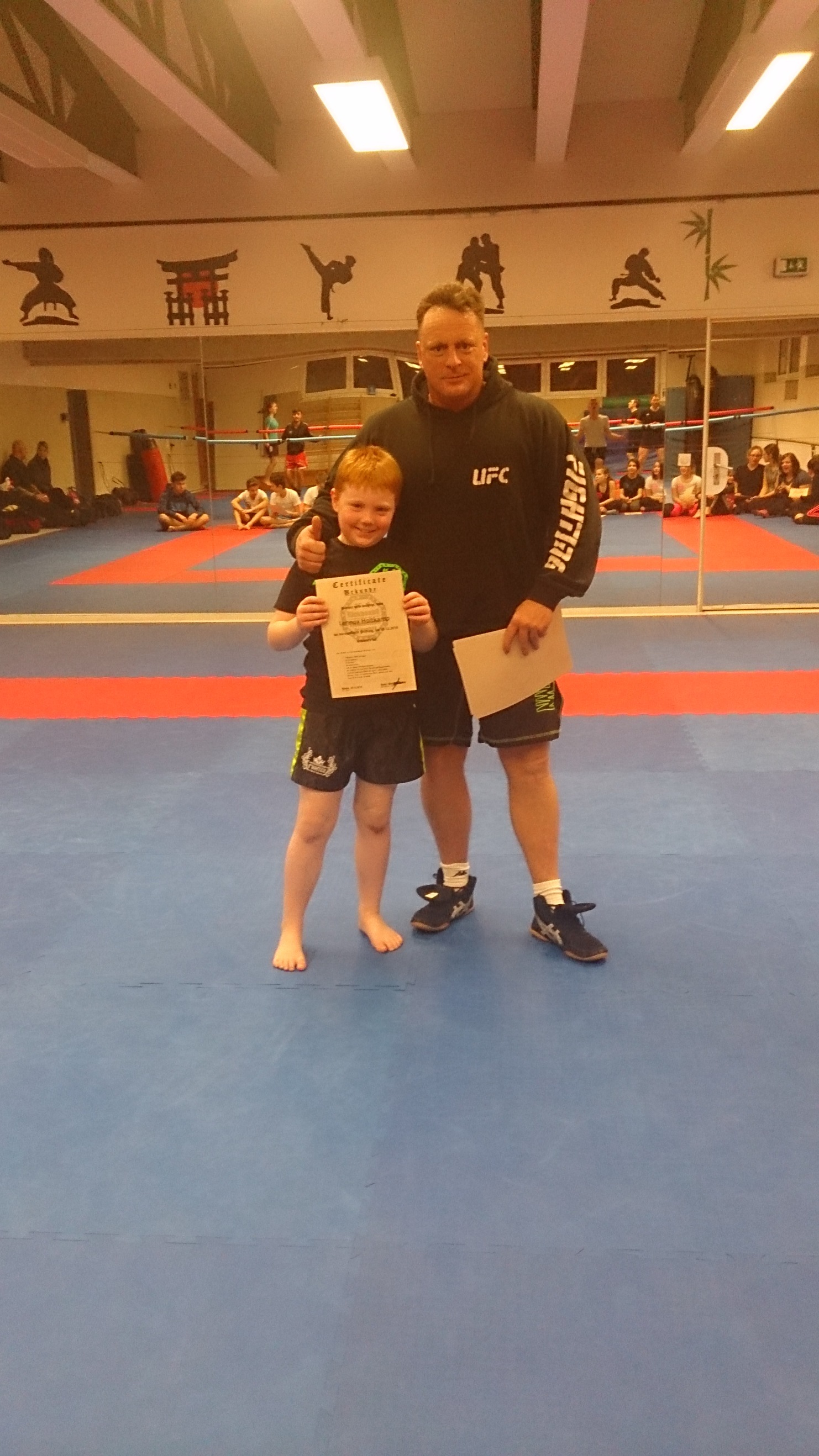 Ein Junge h&auml;lt stolz ein Zertifikat und steht neben einem Trainer in einem Sportbereich mit Matten.