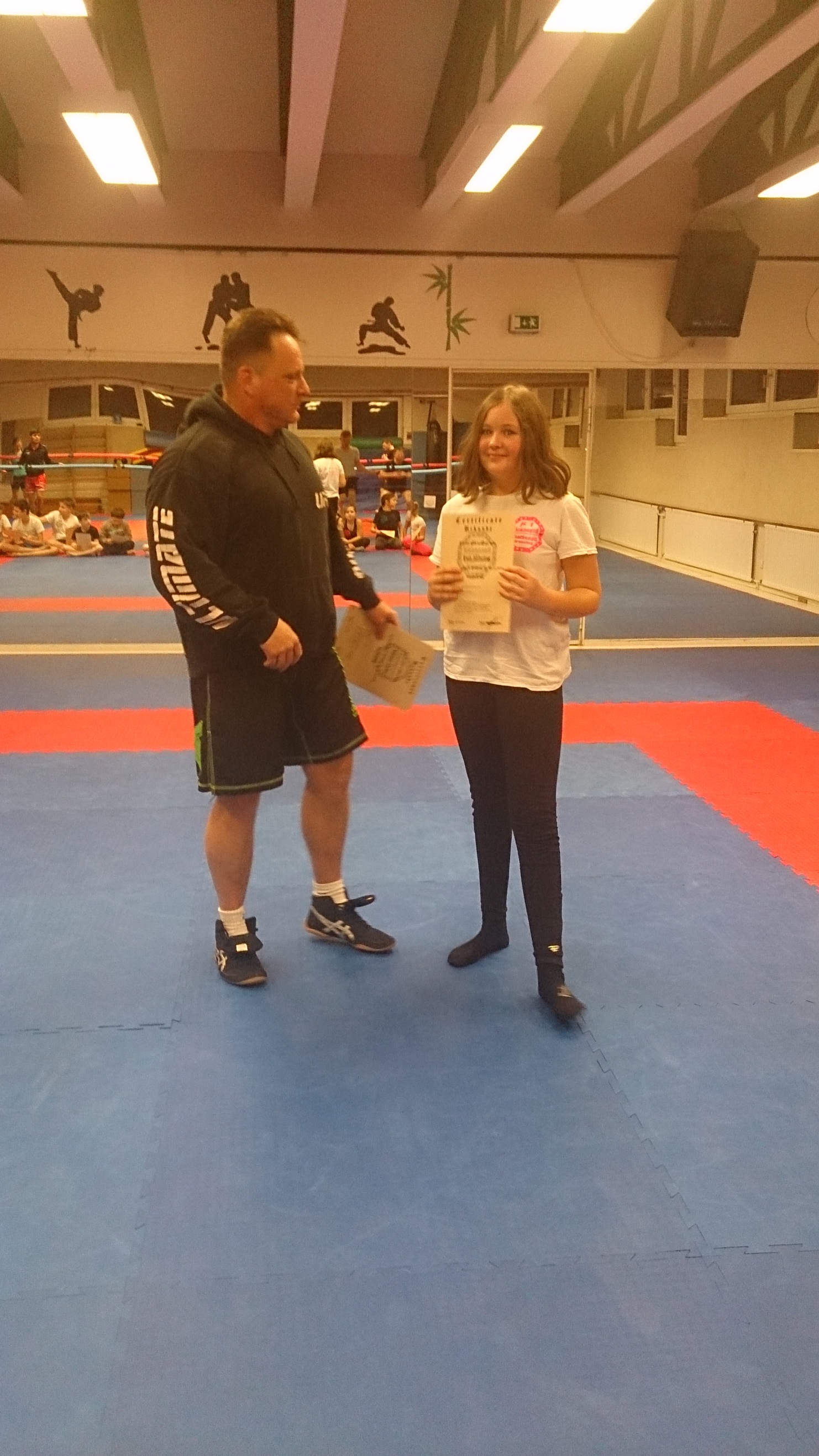 Trainer &uuml;berreicht einer Jugendlichen eine Urkunde in einem Sportstudio mit Teppichfl&auml;che.