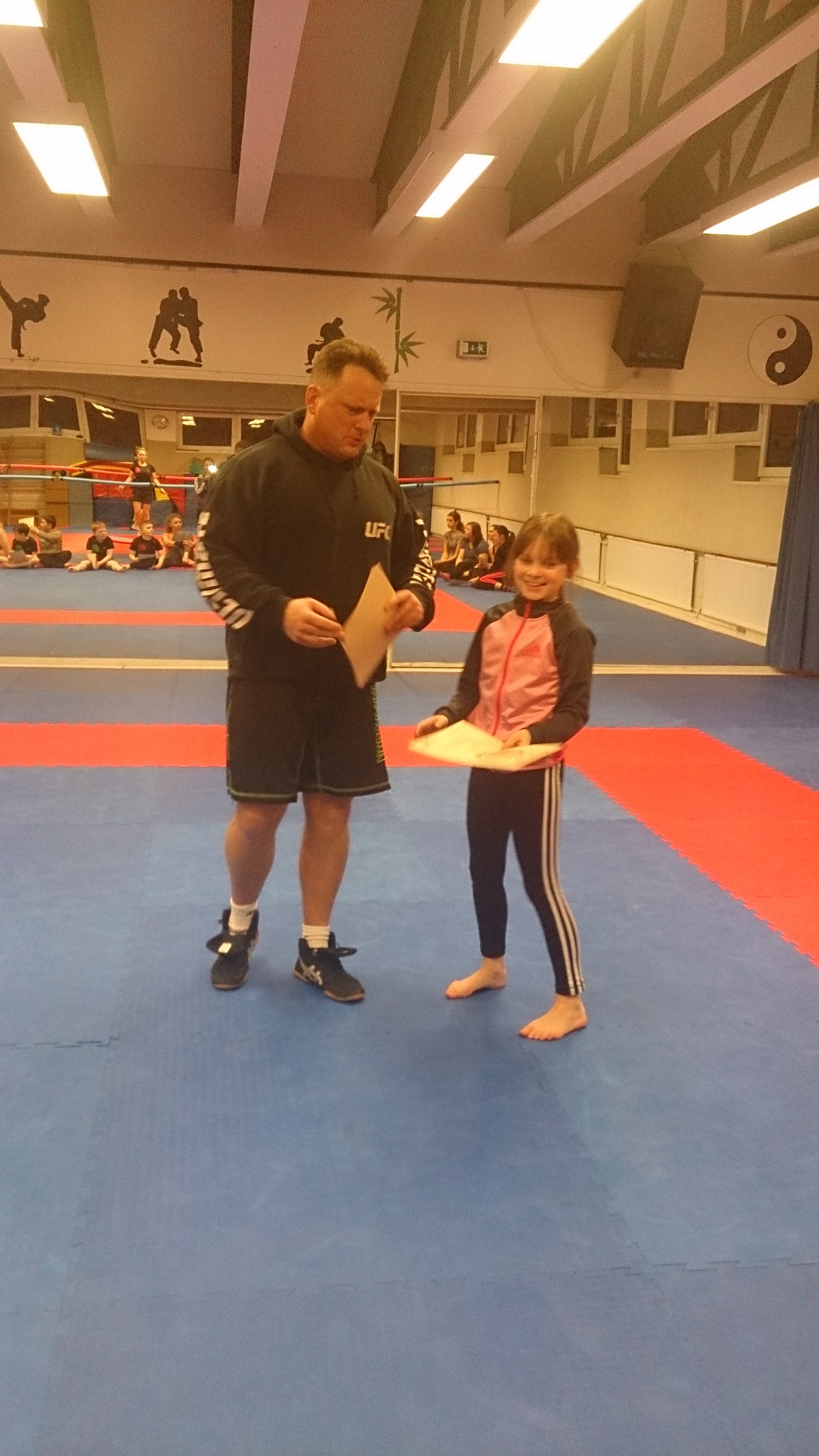 Trainer &uuml;berreicht einem M&auml;dchen eine Urkunde auf einer blauen Trainingsmatte in einem Sportstudio.