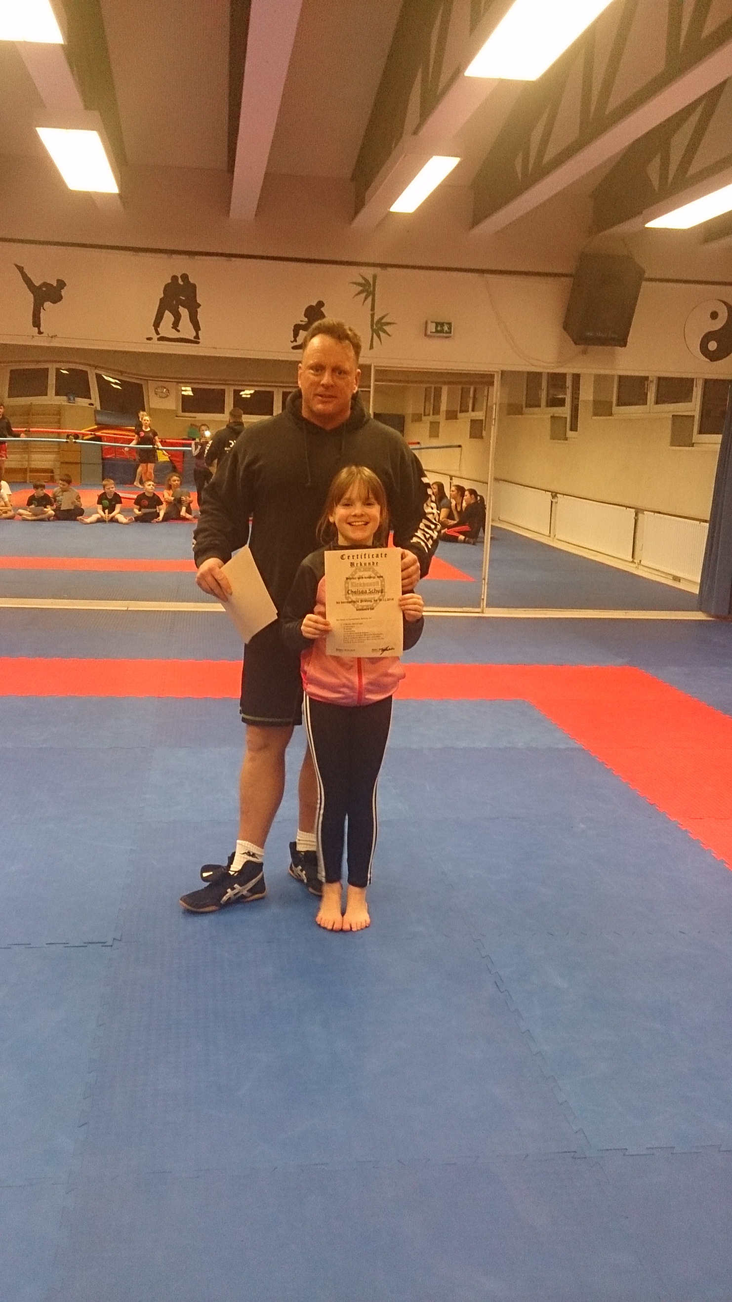 Ein Trainer steht mit einem M&auml;dchen auf einer Trainingsmatte und h&auml;lt ein Zertifikat. Im Hintergrund sind weitere Sportler sichtbar.