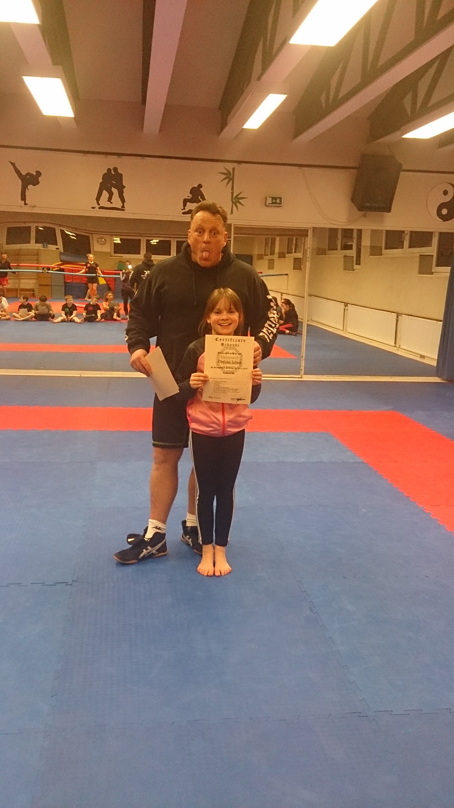 Ein Trainer und ein M&auml;dchen halten ein Zertifikat in einem Kampfsportstudio mit blauer Matte im Hintergrund.