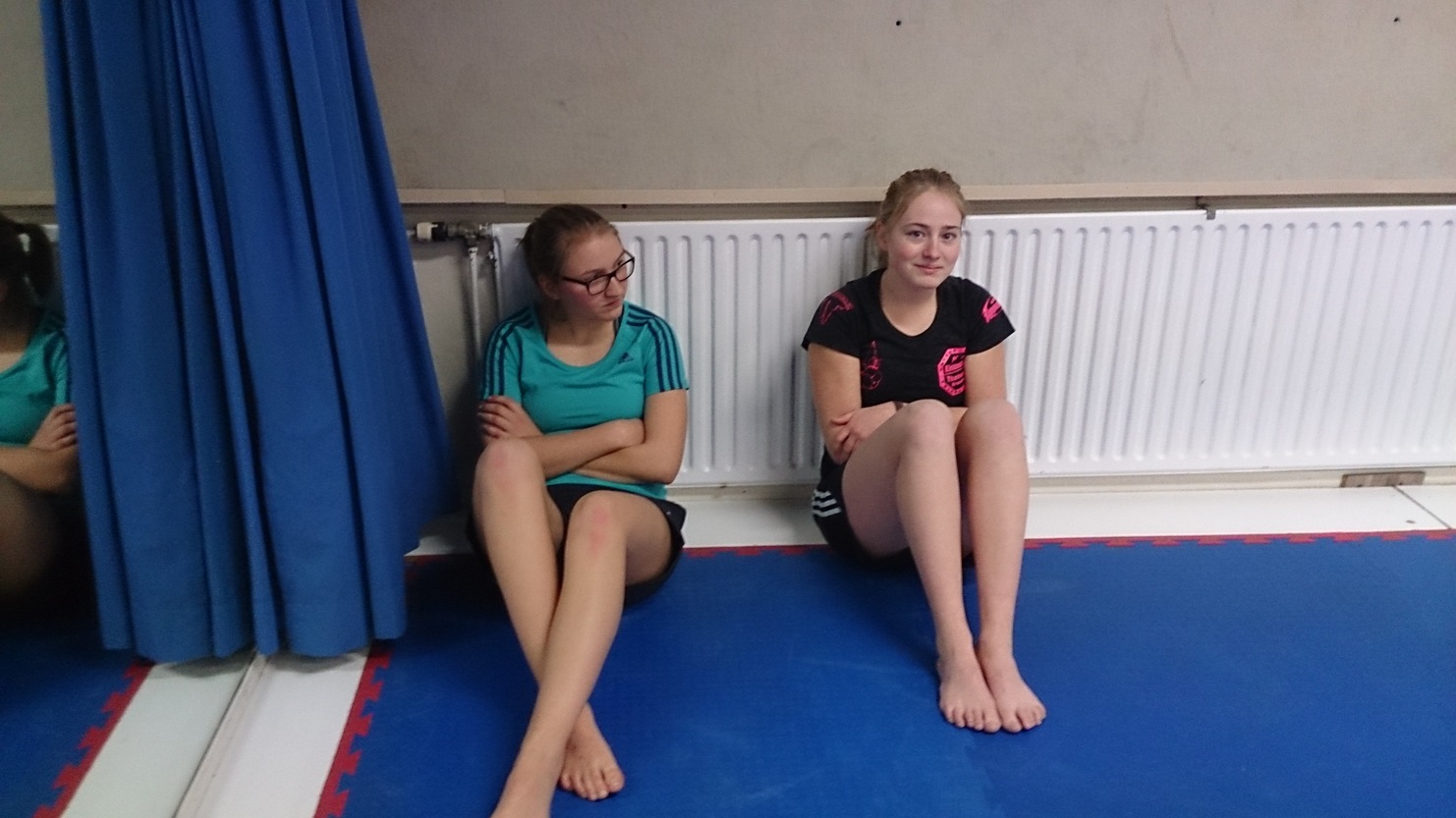 Zwei M&auml;dchen sitzen auf einer blauen Matte an einer Wand, ein Heizk&ouml;rper dahinter, beide in sportlicher Kleidung.