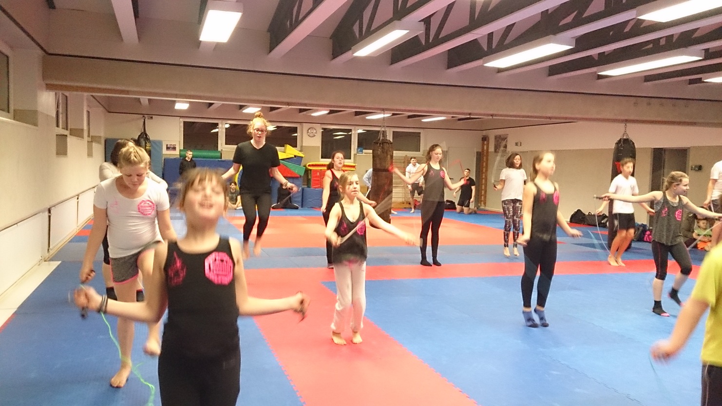 Gruppe von Kindern springt mit Seilen auf blauer und roter Matte in einem Sportraum. Einige tragen Trainingskleidung.