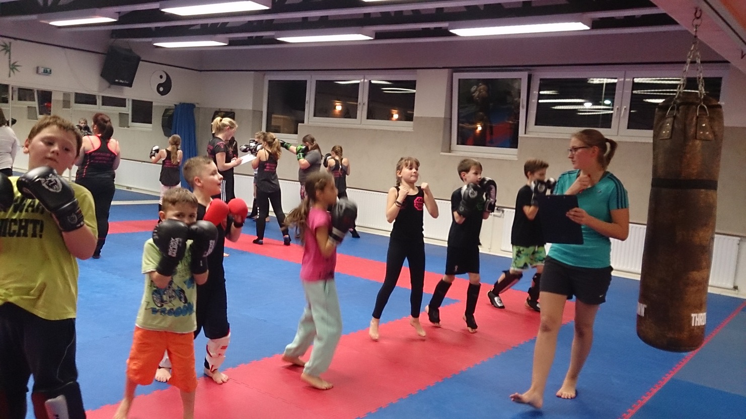 Gruppe von Kindern im Training, die mit Boxhandschuhen auf einer blauen Matte vor einem Sandsack &uuml;ben.