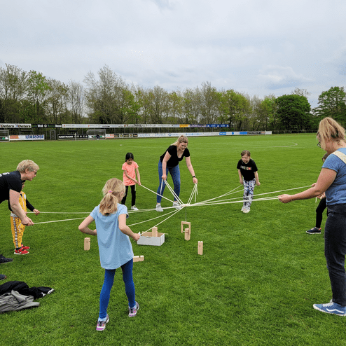 Sechs Kinder und Erwachsene ziehen an Seilen, während sie um Holzklötze auf einem grünen Sportplatz gruppiert sind.