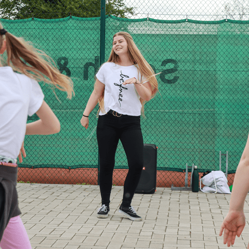 Eine junge Frau in einem weißen T-Shirt tanzt vor einer Gruppe von Mädchen auf einem gepflasterten Platz.