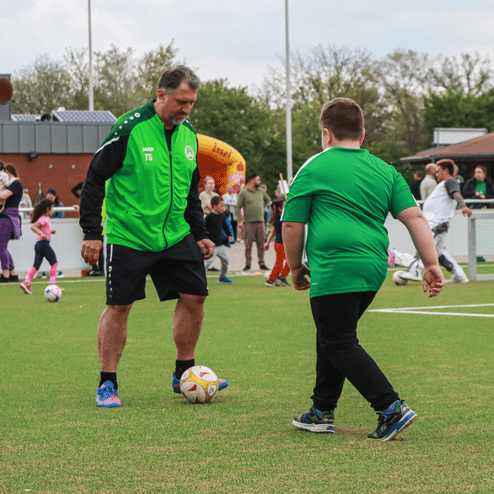 Ein Trainer in grünem Outfit spielt mit einem Jungen in grünem Shirt auf einem Rasenplatz mit Fußball.