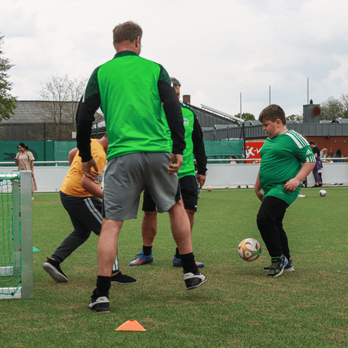 Kinder spielen Fußball auf einem Rasenplatz, während Trainer Anweisungen geben und ein Tor im Vordergrund steht.