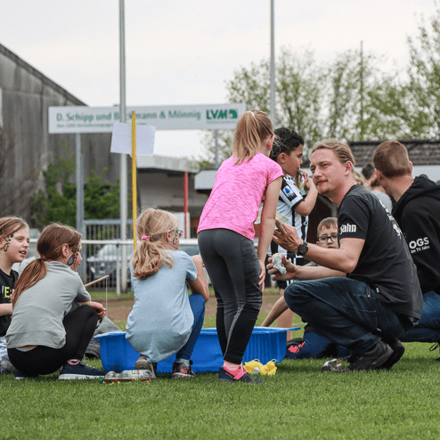 Gruppe von Kindern beim Spiel mit einem Betreuer auf einem Sportplatz, im Hintergrund eine Sporthalle und Bäume.