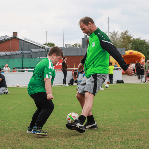 Ein Trainer und ein Junge in grünen Trikots üben das Fußballspielen auf einem Rasenplatz. Zuschauer stehen im Hintergrund.