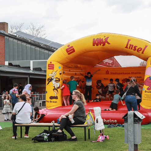 Kinder spielen auf einer bunten Hüpfburg vor einem Gebäude, während andere Zuschauer in der Nähe stehen.