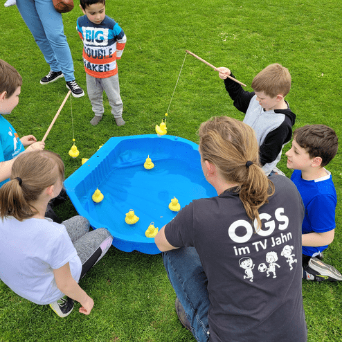Gruppe von Kindern versucht beim Angeln mit Stäben, gelbe Enten aus einem blauen Becken auf einer Wiese zu fangen.