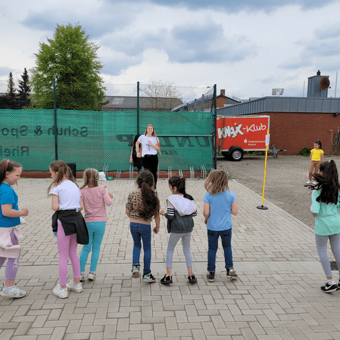 Gruppe von Kindern steht mit dem Rücken zur Kamera vor einer Sporteinheit, Trainerin spricht mit ihnen auf einem Gelände.