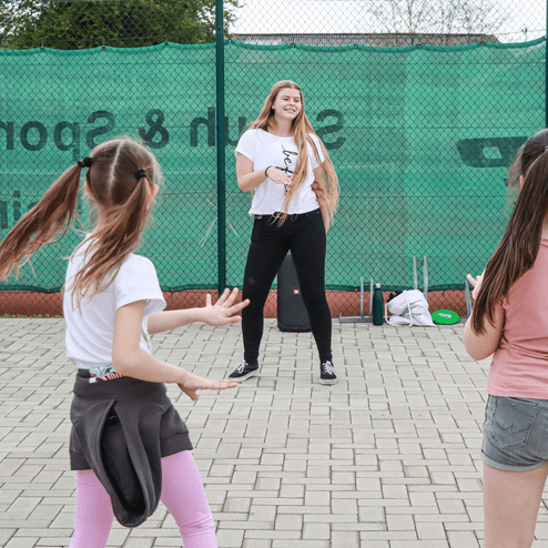 Trainerin leitet Kinder in einer Sportübung auf einem Platz vor einem Netz mit grüner Abdeckung an.
