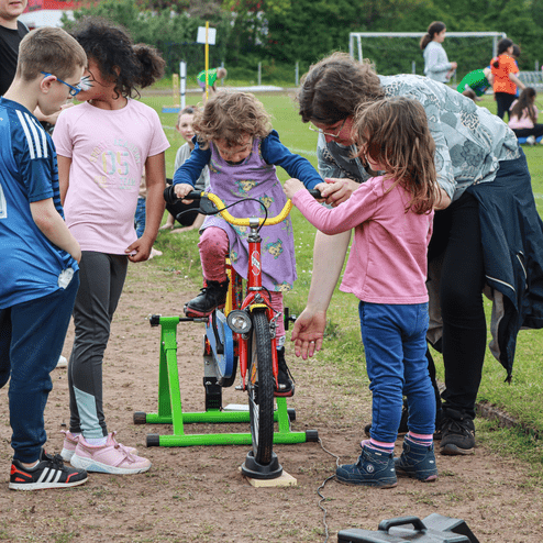 Gruppe von Kindern beobachtet eine Frau, die einem Mädchen auf einem Fahrrad hilft, auf einem Sportplatz.