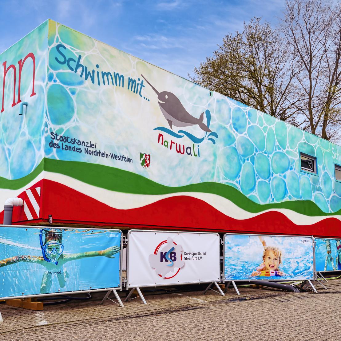 Bunt gestalteter Anh&auml;nger mit Motiven von Wasser und einem Narwal, umgeben von Sichtschutzw&auml;nden mit Schwimm-Themen.