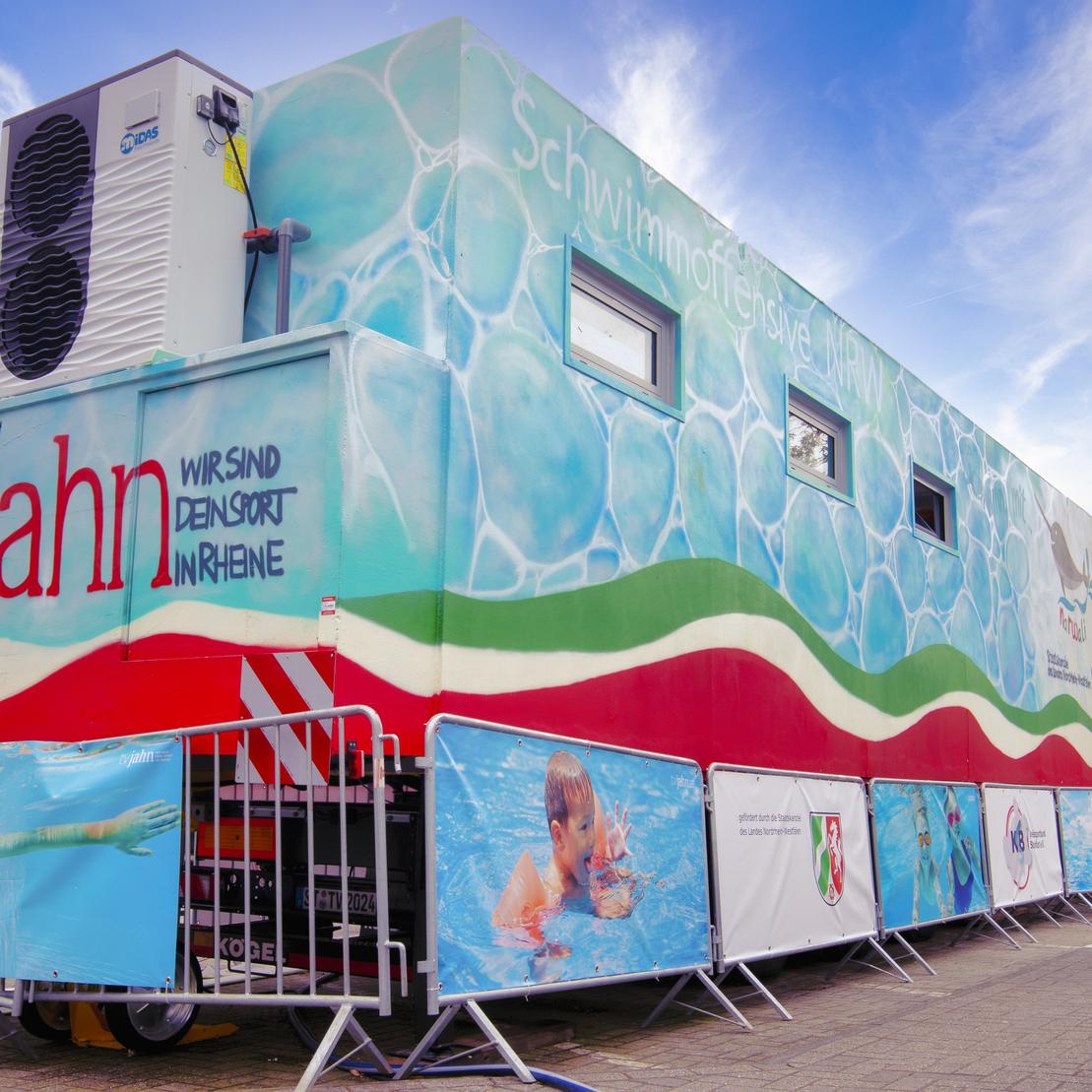 Mobiles Schwimmbad mit bunter Wandbemalung, umgeben von Absperrungen und Werbebannern auf einem Au&szlig;enbereich.