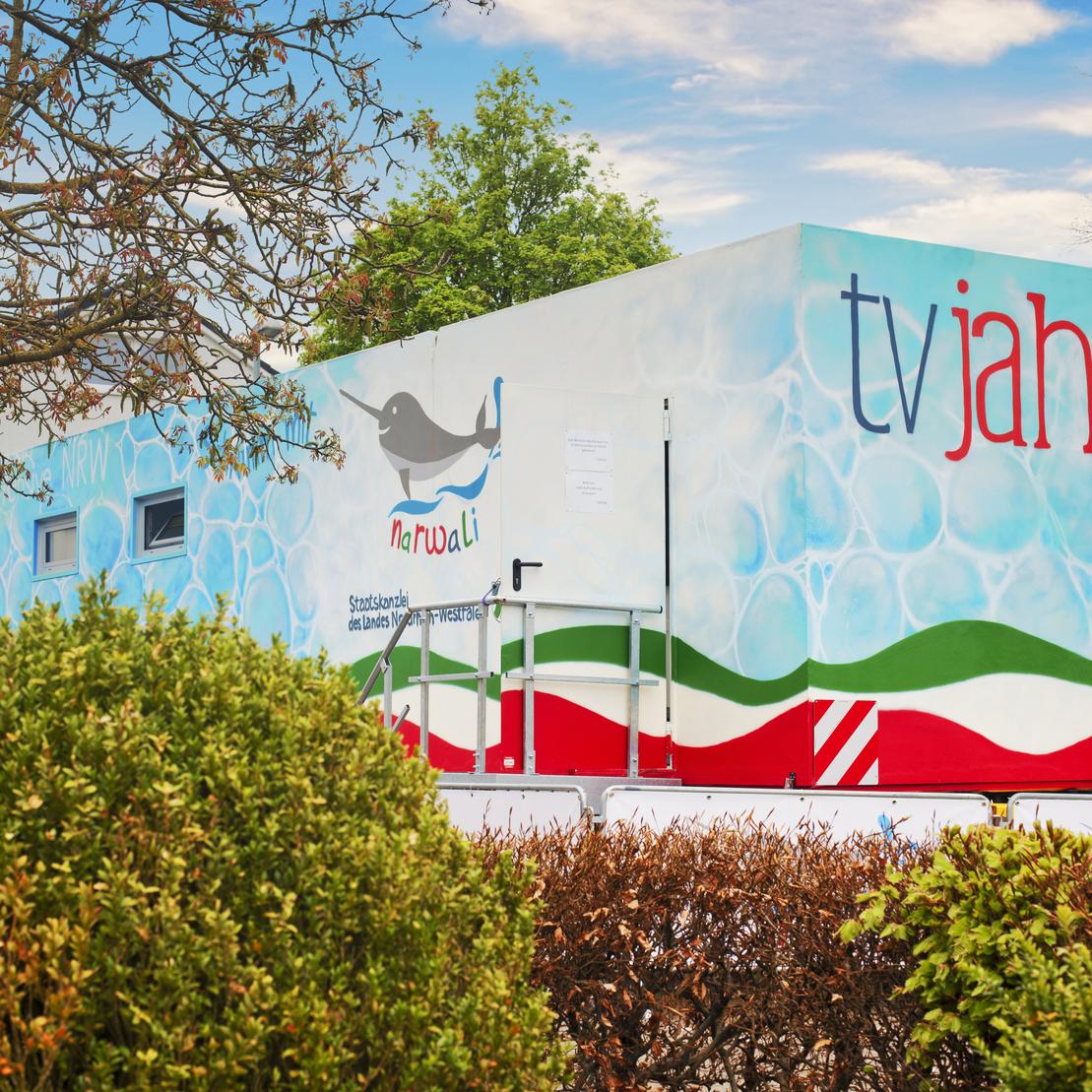 Bunte mobile Verkaufsstelle mit der Aufschrift &bdquo;tv jahn&ldquo;, umgeben von gr&uuml;nen B&uuml;schen und B&auml;umen.