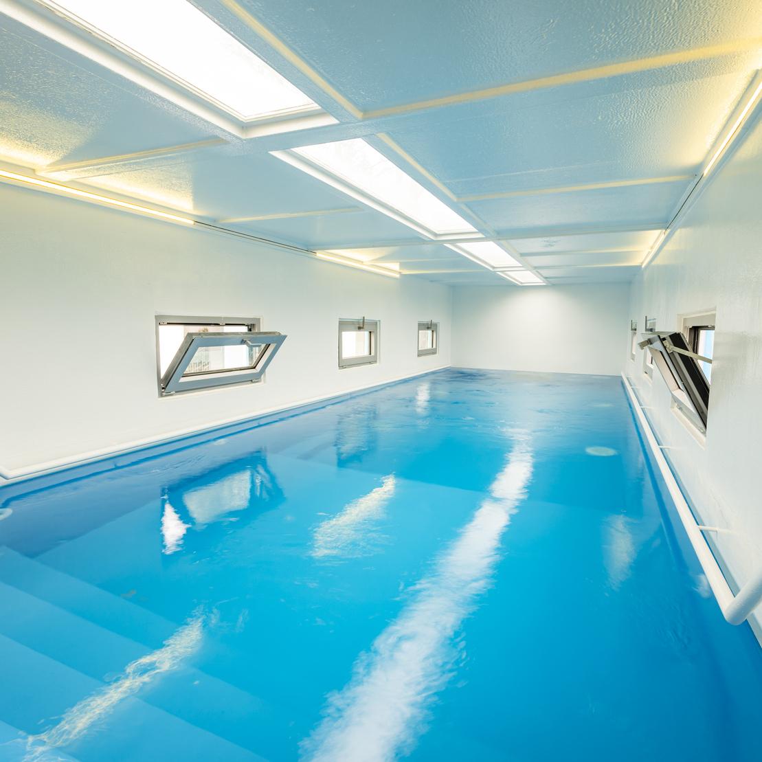 Blick in einen hellen Raum mit blauer Wasserfl&auml;che und Fenstern, die Tageslicht hereinlassen.