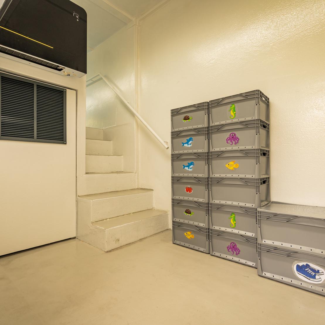 Innenraum mit einer Treppe, einer geschlossenen T&uuml;r und einem Stapel bunter Schuhboxen aus Kunststoff.