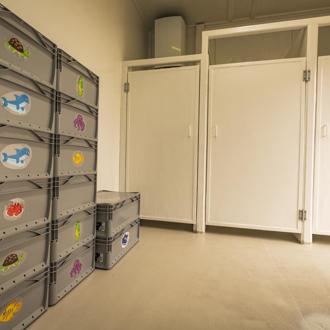 Stapelhaufen aus grauen Kunststoffkisten mit bunten Stickern, neben wei&szlig;en Trennw&auml;nden in einem Raum.