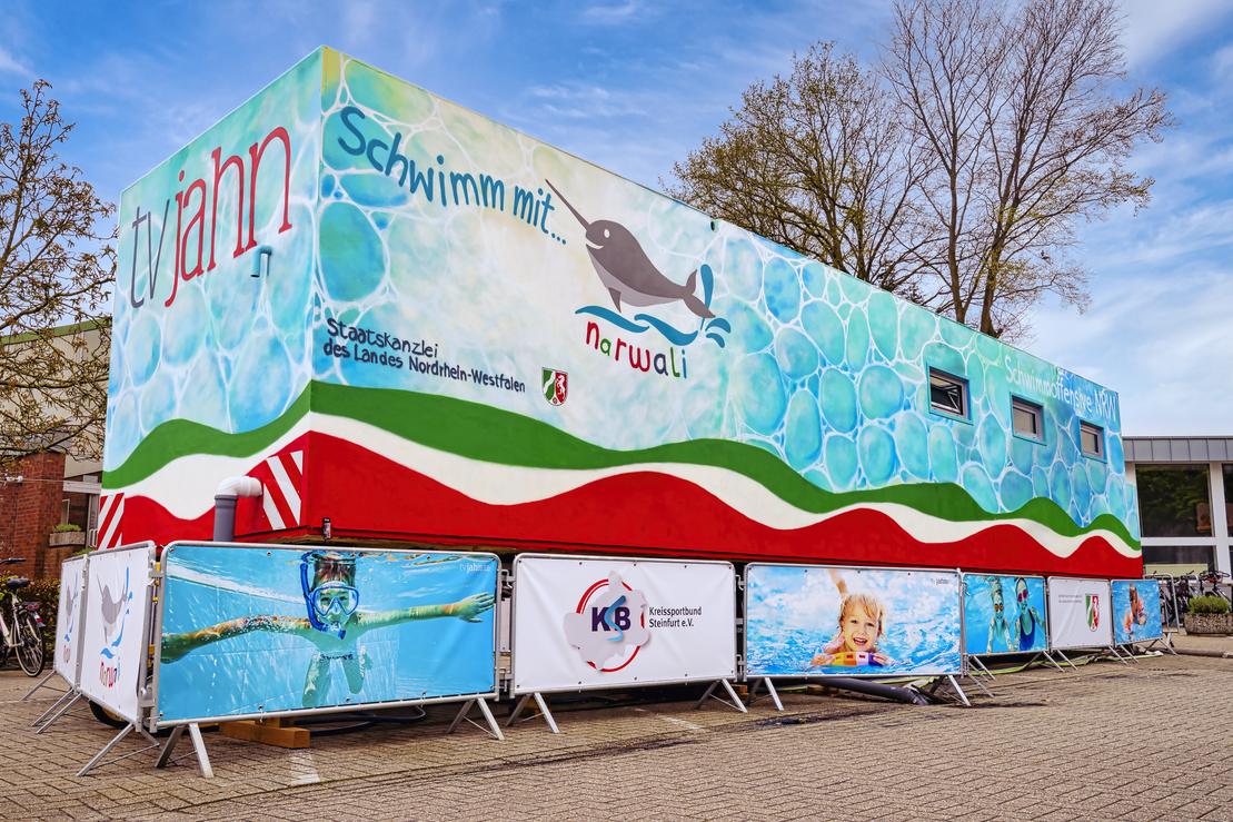 Bunt gestalteter Anh&auml;nger mit Motiven von Wasser und einem Narwal, umgeben von Sichtschutzw&auml;nden mit Schwimm-Themen.