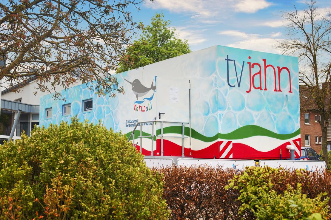 Bunte mobile Verkaufsstelle mit der Aufschrift &bdquo;tv jahn&ldquo;, umgeben von gr&uuml;nen B&uuml;schen und B&auml;umen.