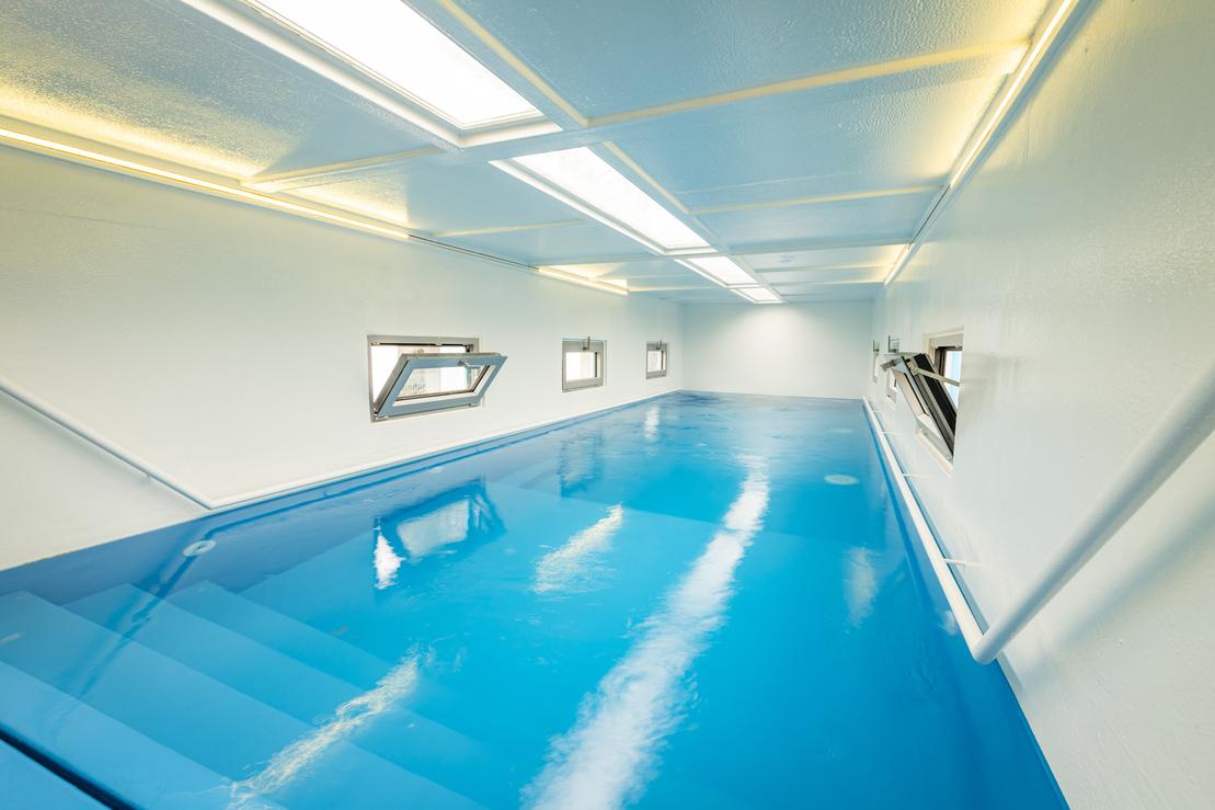 Blick in einen hellen Raum mit blauer Wasserfl&auml;che und Fenstern, die Tageslicht hereinlassen.