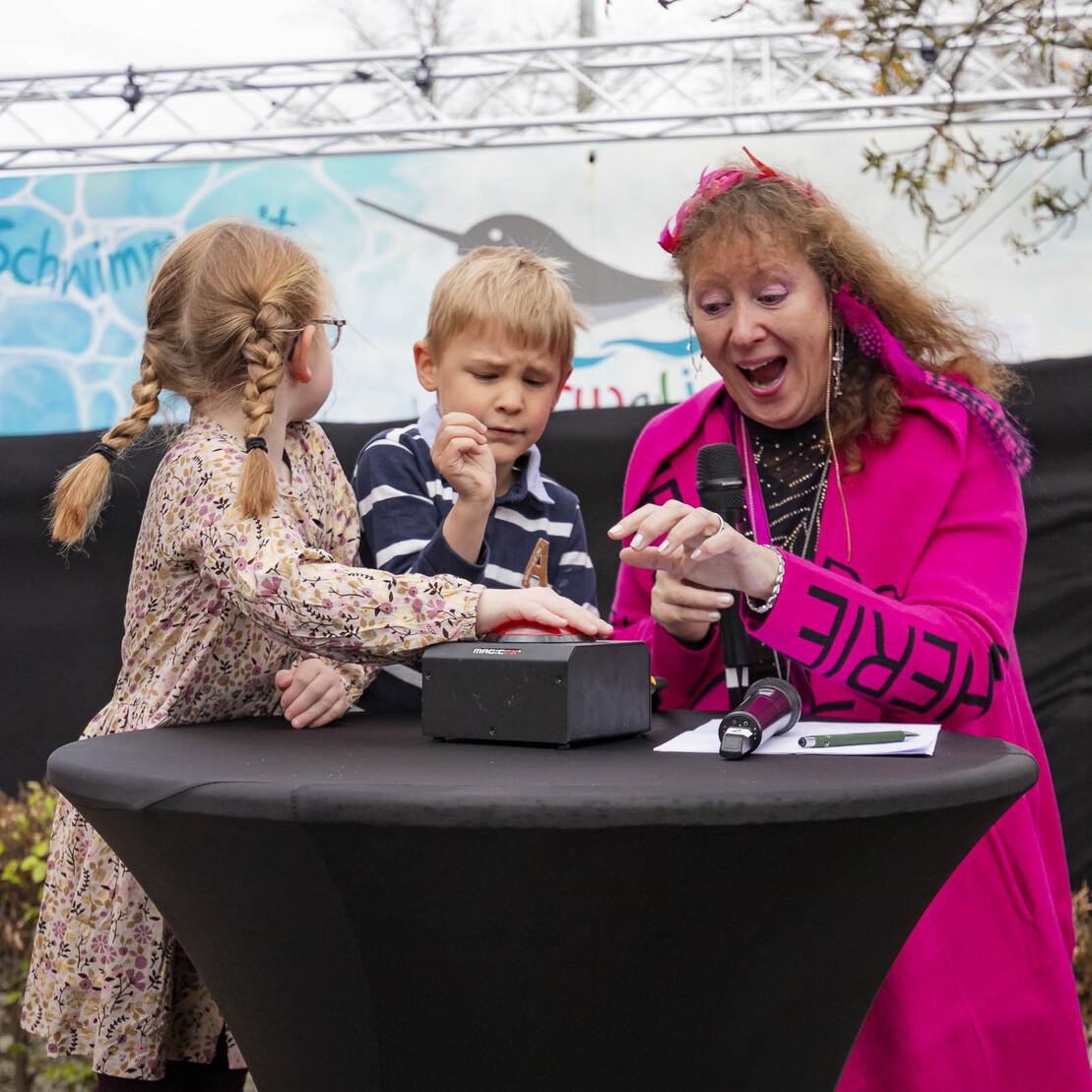 Drei Personen stehen an einem Tisch, eine Frau in pinkem Outfit erkl&auml;rt Kindern mit Spa&szlig; ein Spielger&auml;t.