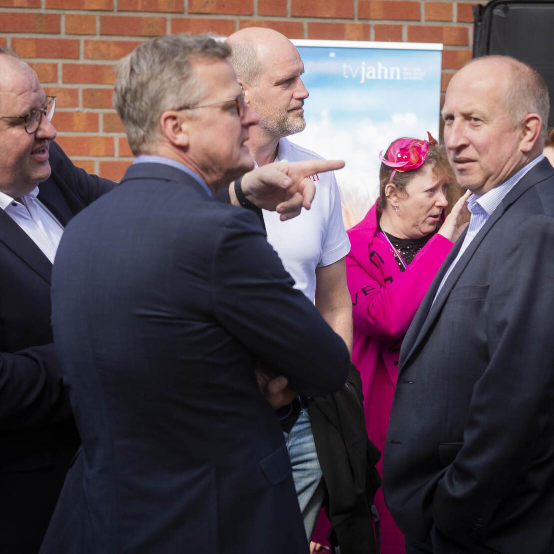 F&uuml;nf Personen stehen in einer Gruppe, w&auml;hrend sie miteinander sprechen und l&auml;cheln, vor einer Backsteinmauer.