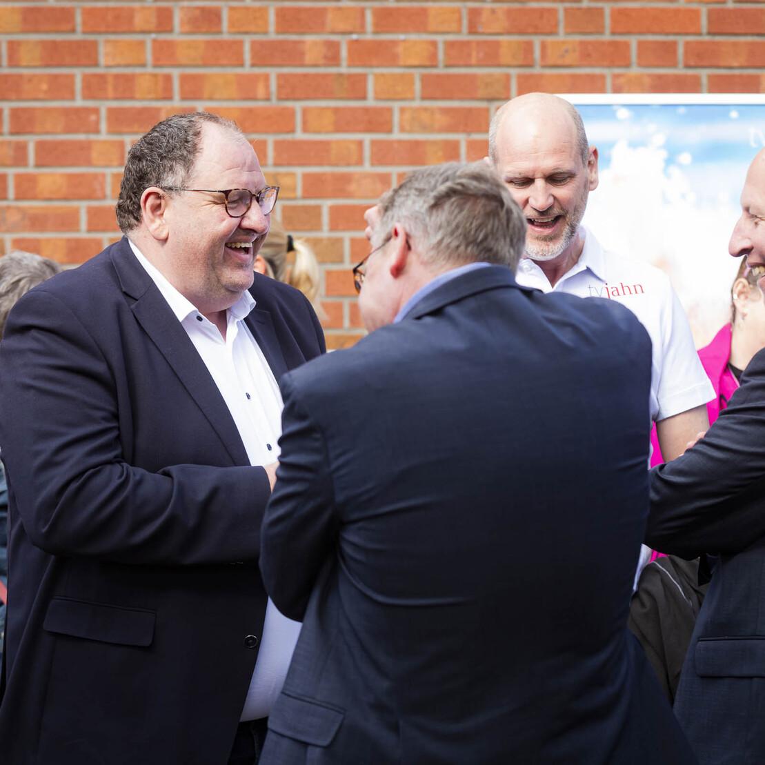 Vier M&auml;nner unterhalten sich l&auml;chelnd im Freien, w&auml;hrend einige Personen im Hintergrund stehen. Ziegelwand sichtbar.