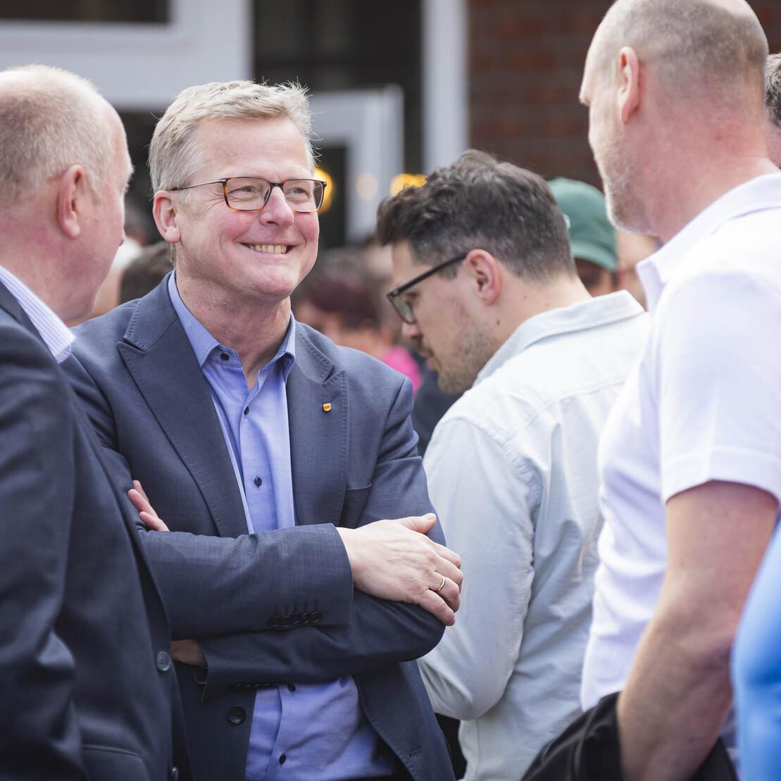 Gruppe von Personen im Gespr&auml;ch, mit einem l&auml;chelnden Mann in Anzug und Brille im Vordergrund.