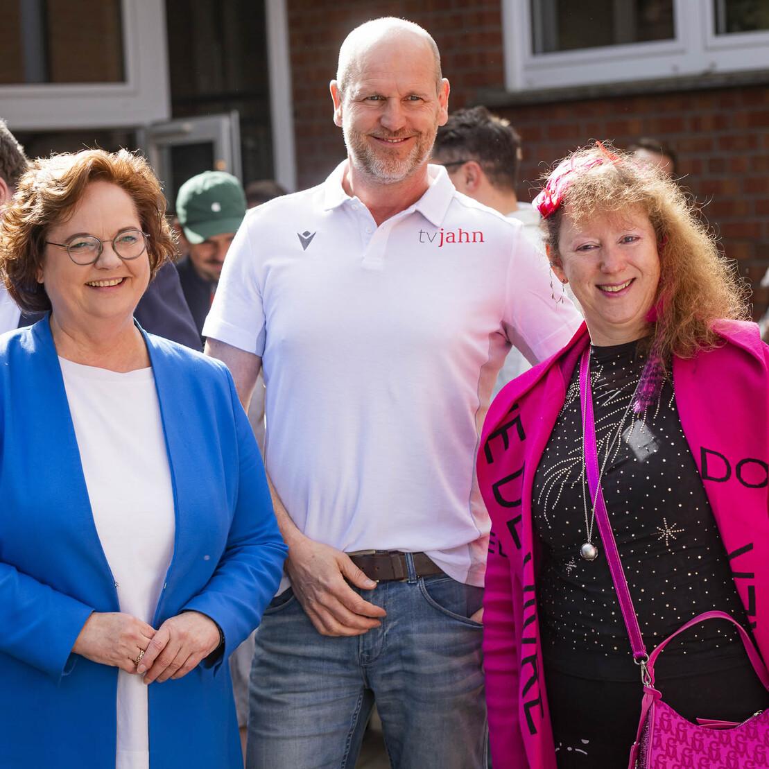 Drei Personen stehen l&auml;chelnd vor einer Gruppe von Menschen in einem Garten, umgeben von gr&uuml;nen Pflanzen und Geb&auml;uden.