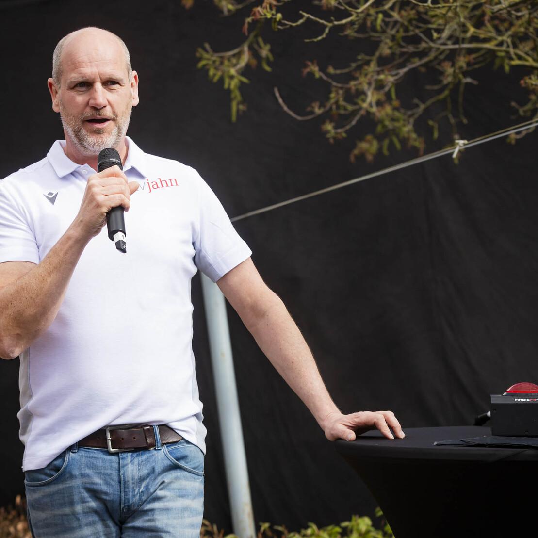 Mann mit Mikrofon spricht an einem Podium vor schwarzem Hintergrund im Freien.
