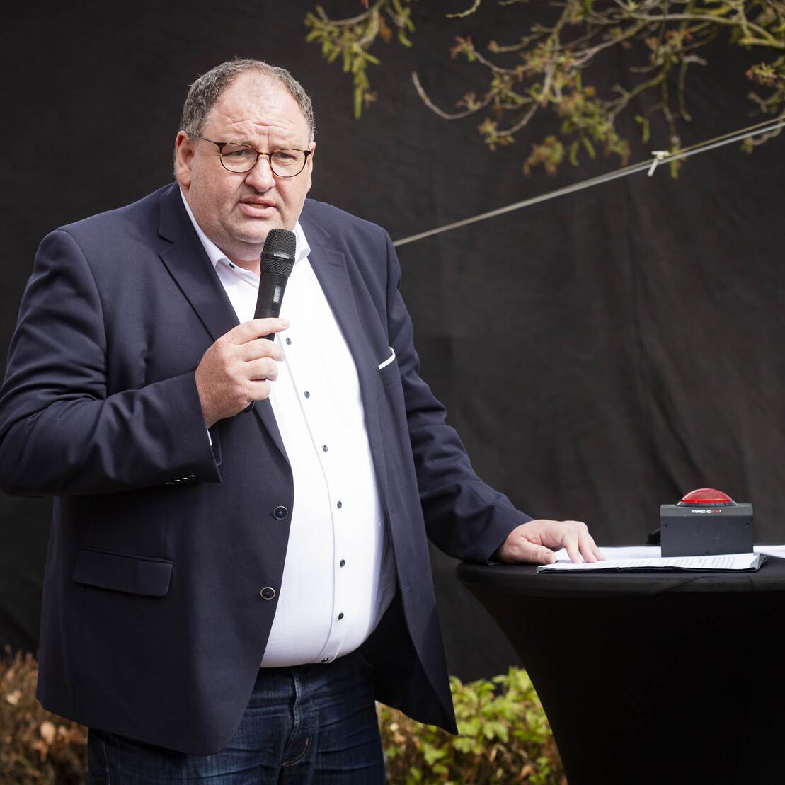 Mann mit Mikrofon spricht an einem Stehtisch im Freien, untermalt von einer dunklen Hintergrundkulisse.