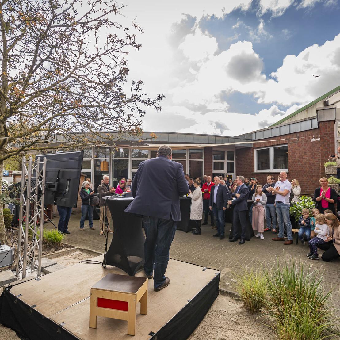 Person h&auml;lt eine Rede vor einer Gruppe von Zuh&ouml;rern im Freien, umgeben von einem Baum und einem Geb&auml;ude.