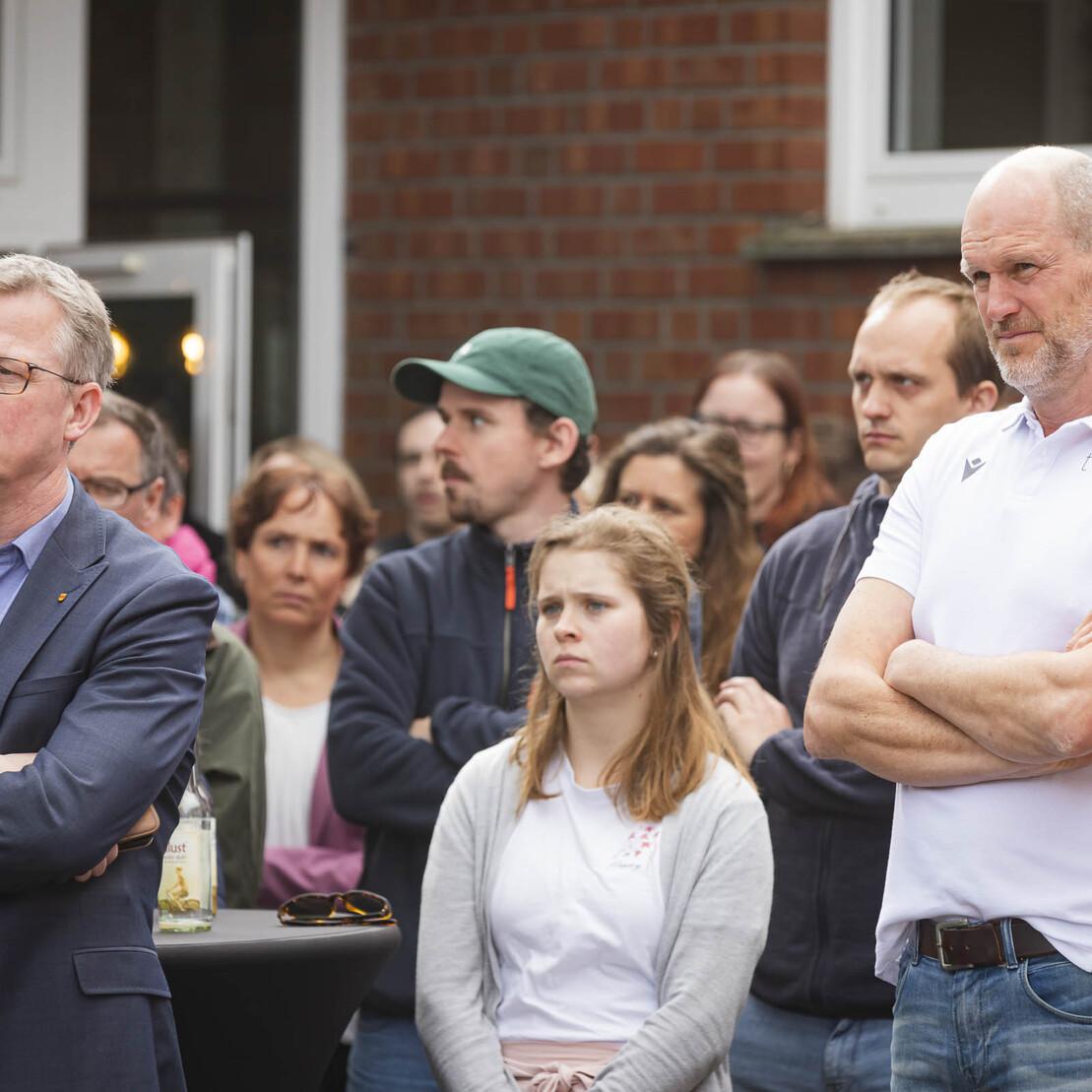 Gruppenszene mit mehreren Personen, die aufmerksam einer Ansprache folgen, einige mit verschr&auml;nkten Armen.