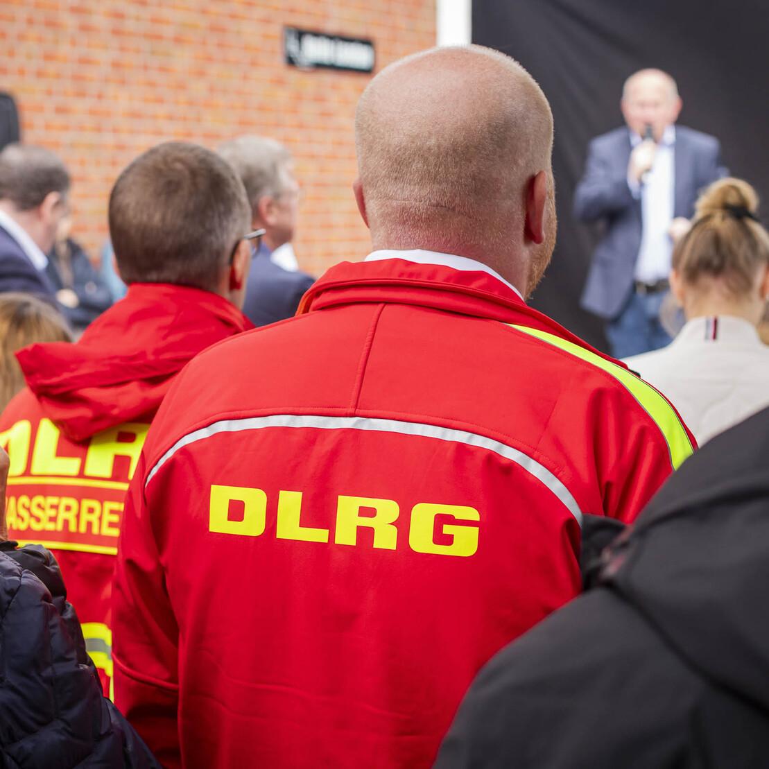 Menschen stehen im Hintergrund, w&auml;hrend ein Redner auf einer B&uuml;hne spricht, DLRG-Uniformen sind deutlich sichtbar.
