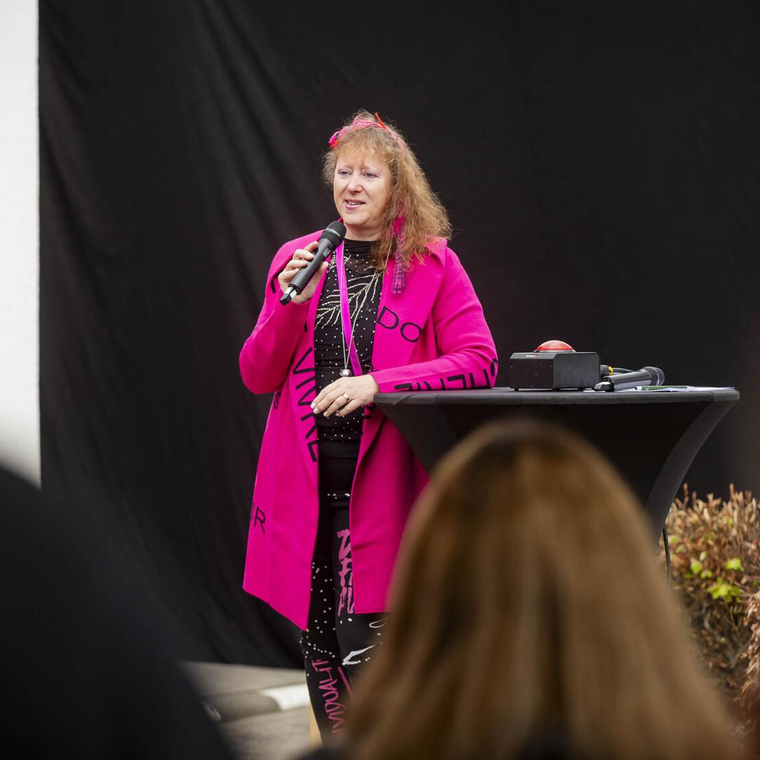 Frau mit langen, lockigen Haaren spricht am Mikrofon in knallpinkem Outfit vor einer Gruppe von Zuh&ouml;rern.