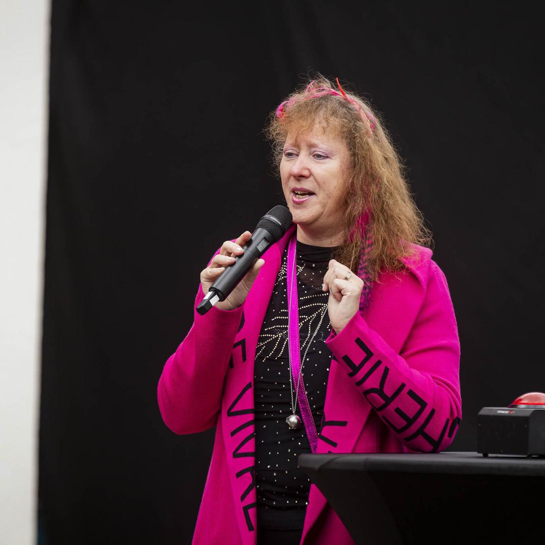 Frau mit Mikrofon spricht lebhaft, tr&auml;gt einen pinken Blazer und steht vor schwarzem Hintergrund auf einer Veranstaltungsb&uuml;hne.