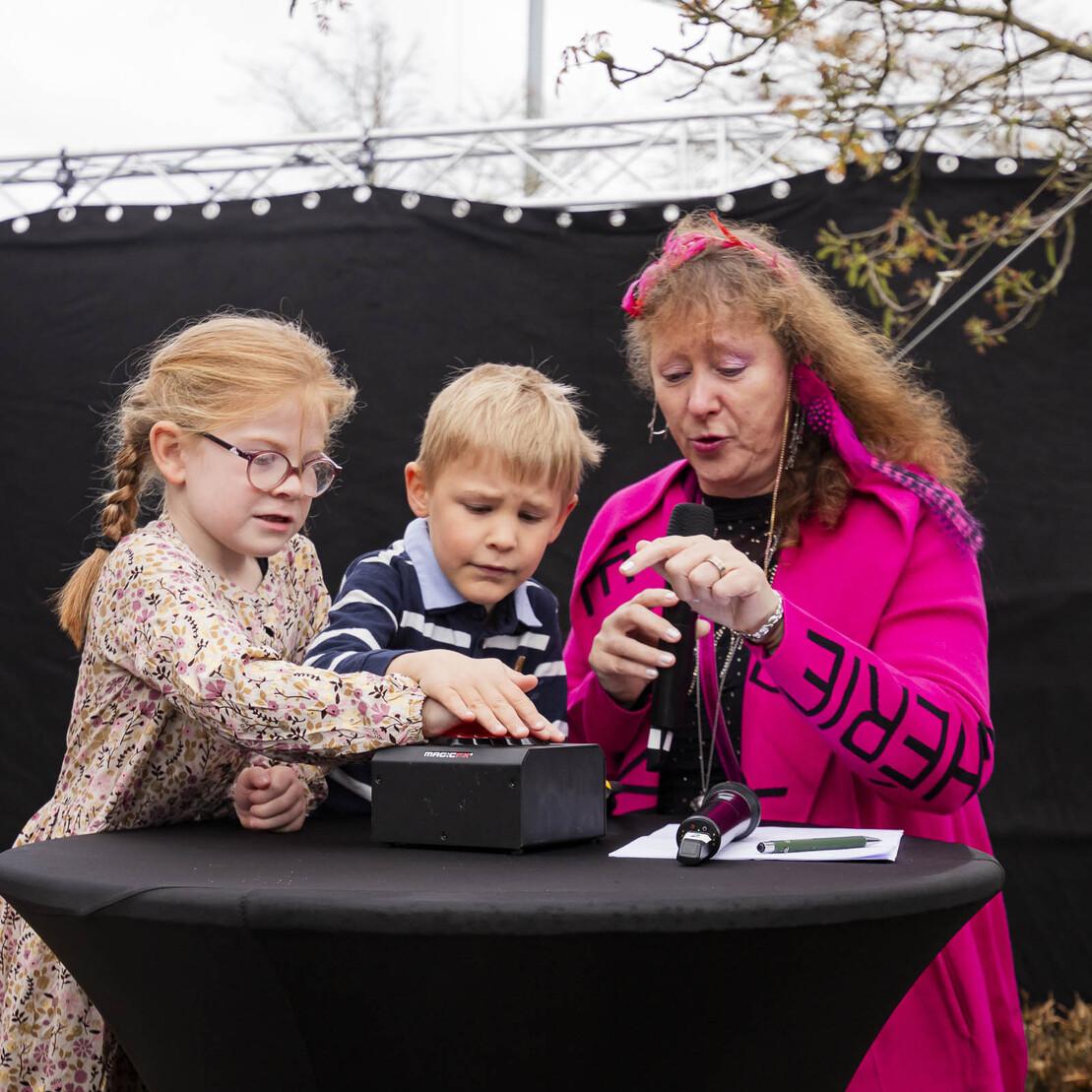 Zwei Kinder und eine Frau interagieren an einem Tisch mit einer Box, umgeben von einem schwarzen Hintergrund.