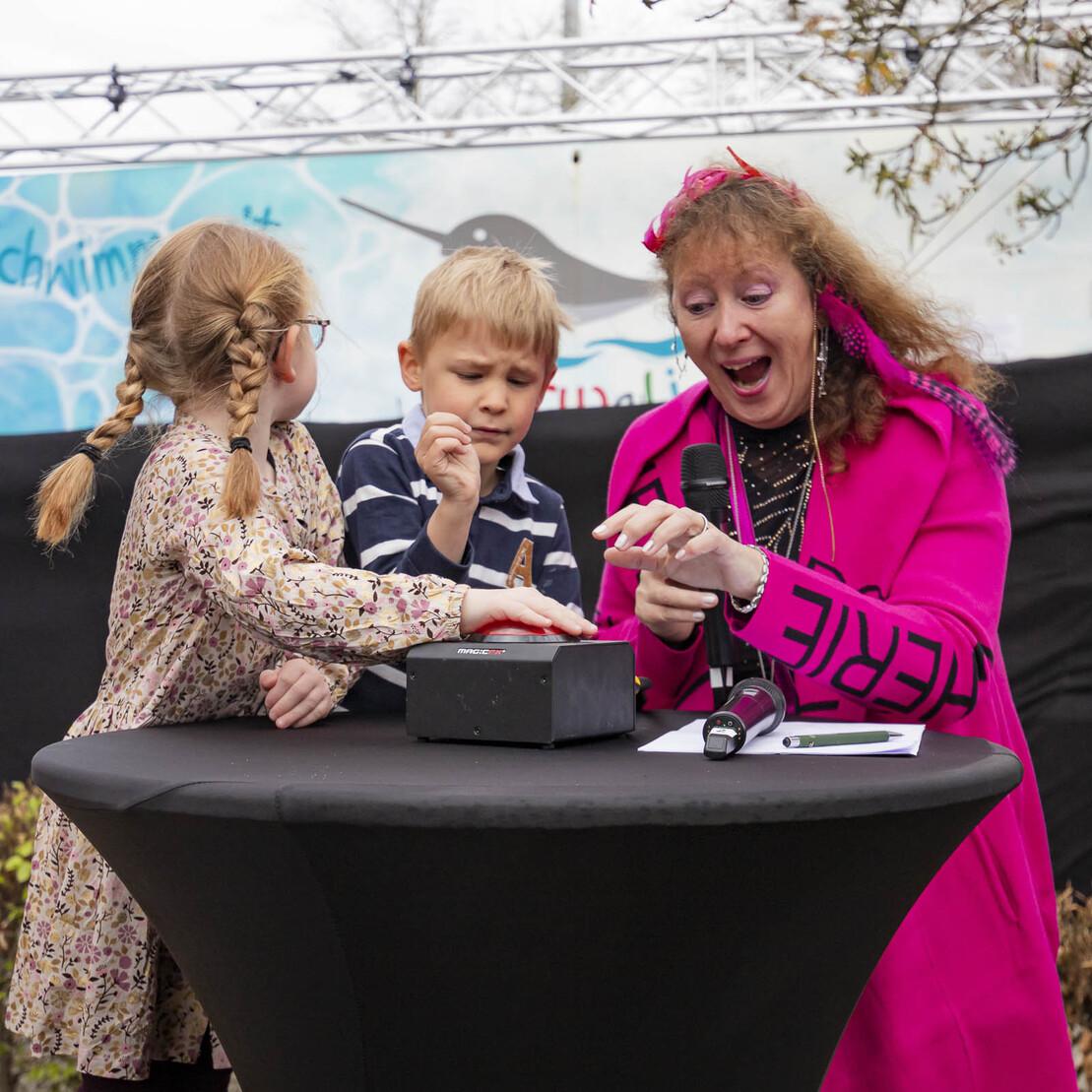 Drei Personen stehen an einem Tisch, eine Frau in pinkem Outfit erkl&auml;rt Kindern mit Spa&szlig; ein Spielger&auml;t.
