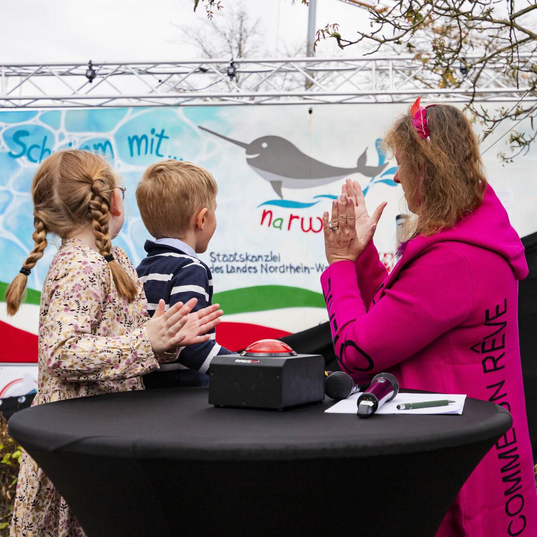 Zwei Kinder und eine Frau in pinkem Outfit stehen an einem Tisch, im Hintergrund ein buntes Werbemotiv.
