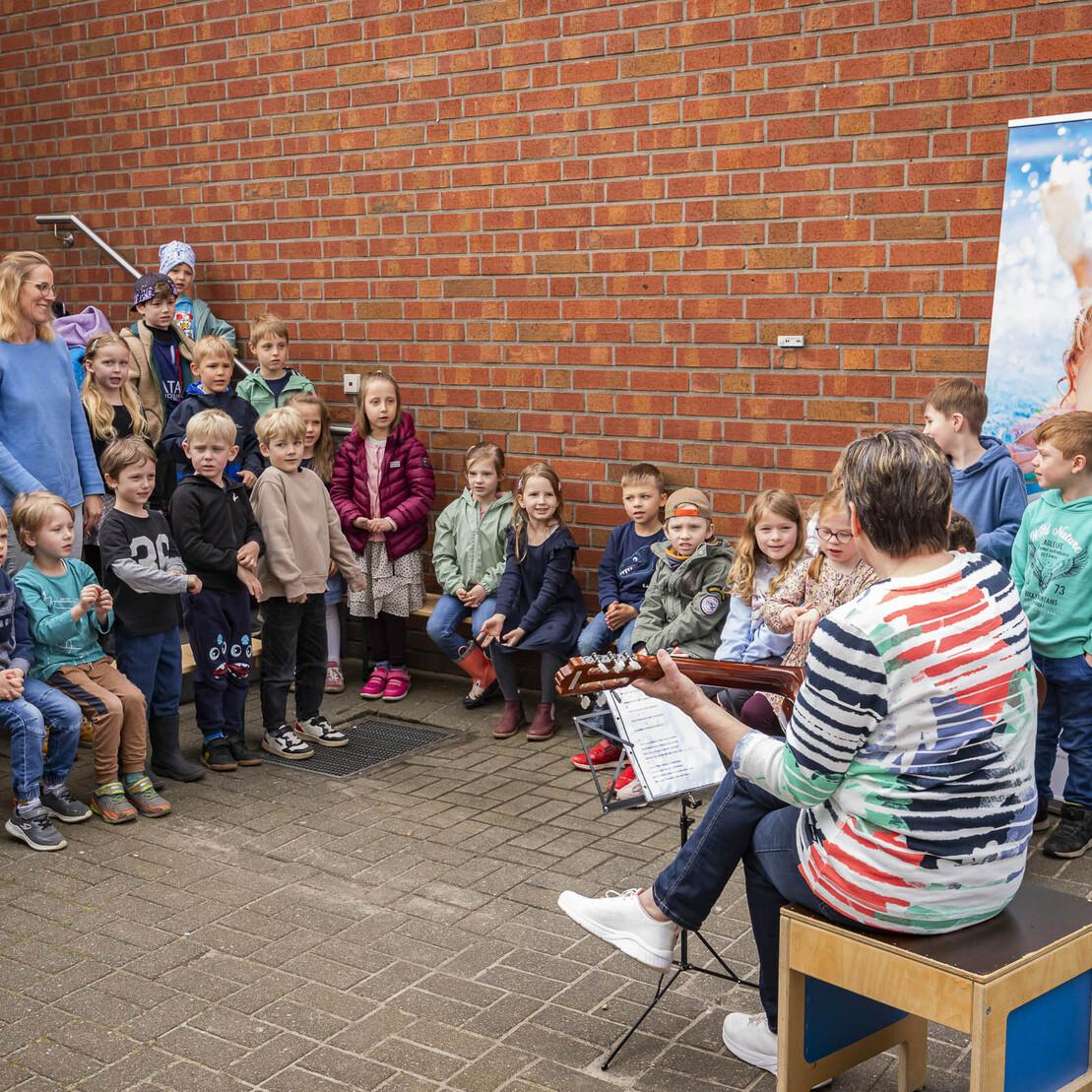 Eine Gruppe von Kindern h&ouml;rt einer Frau mit Gitarre w&auml;hrend einer musikalischen Veranstaltung im Freien zu.