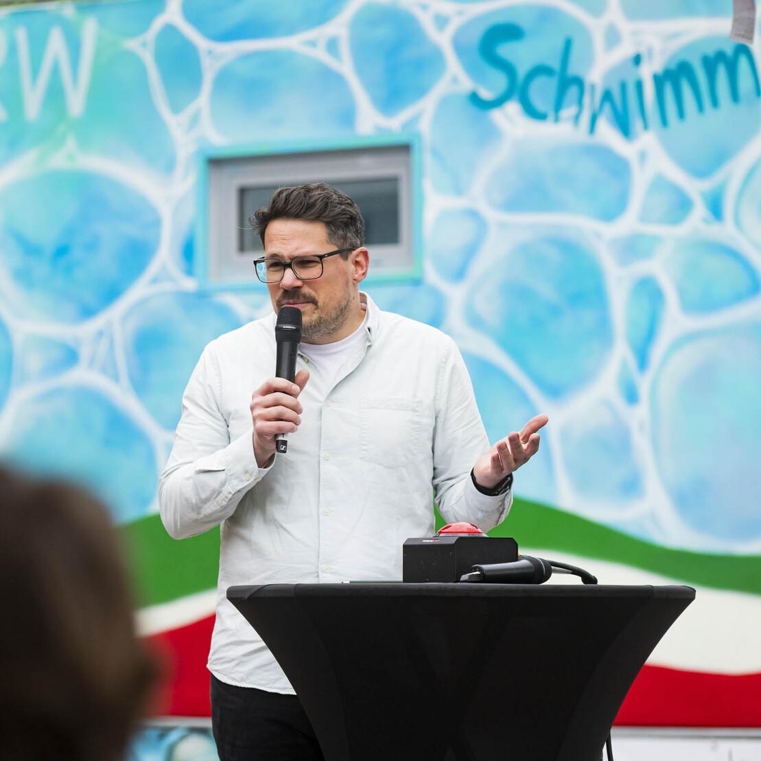 Mann spricht am Mikrofon vor einer muralbemalten Wand mit blauen und gr&uuml;nen Mustern.