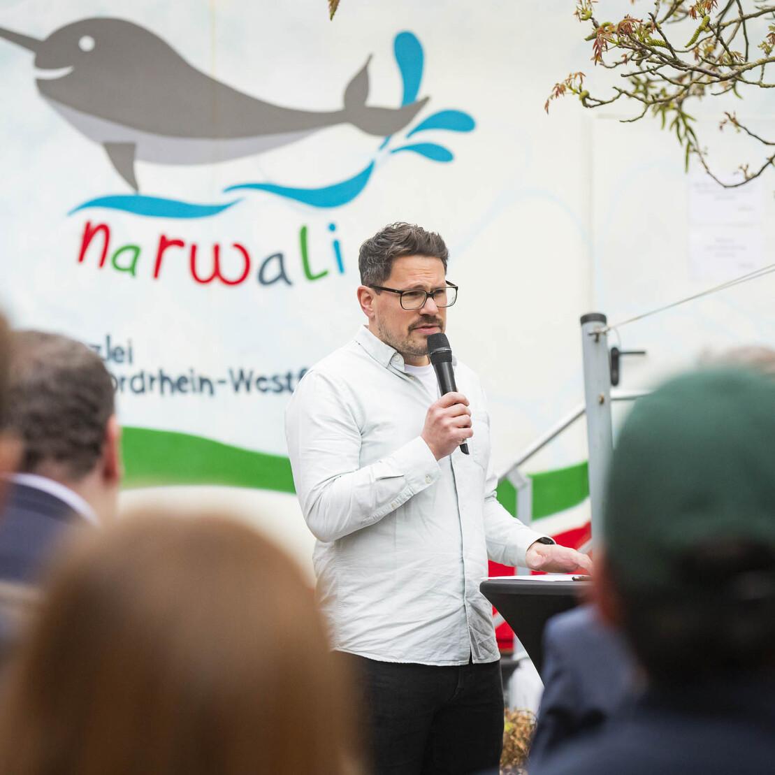 Mann mit Mikrofon spricht vor einer Gruppe von Menschen vor einer muralen Darstellung eines Narwals.