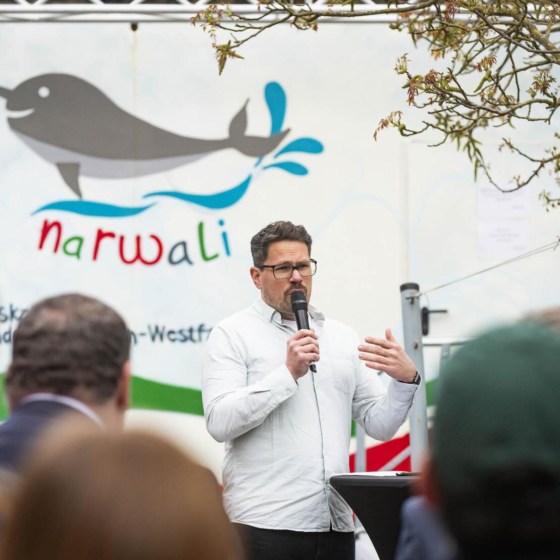 Mann mit Mikrofon spricht vor bunt gestalteter Wand mit Narwal-Motiv und Schriftzug &bdquo;narwali&ldquo;. Menschen in der Zuschauermenge.