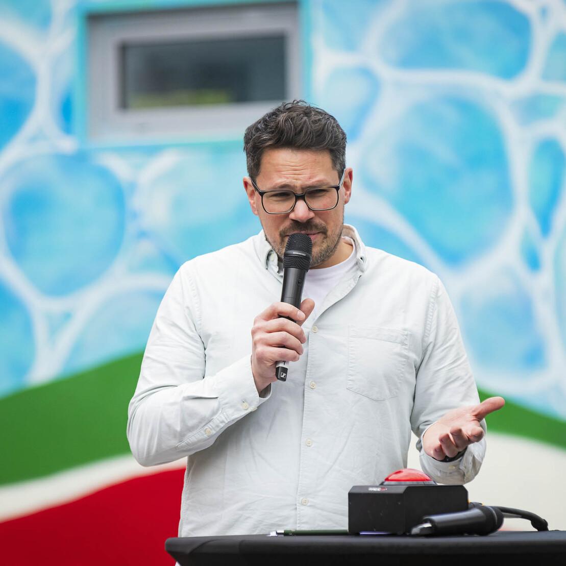 Mann mit Brille spricht mit Mikrofon vor einer bunten Wand aus blauen und gr&uuml;nen Farben.