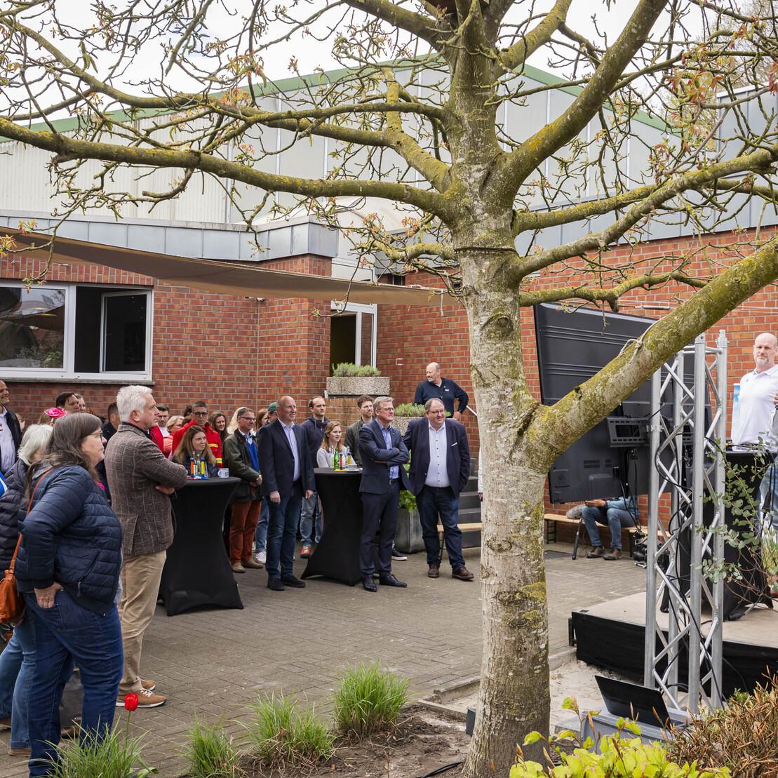 Menschenmenge im Freien lauscht einem Redner auf einer B&uuml;hne zwischen B&auml;umen und Geb&auml;uden. Gr&uuml;ne Pflanzen im Vordergrund.