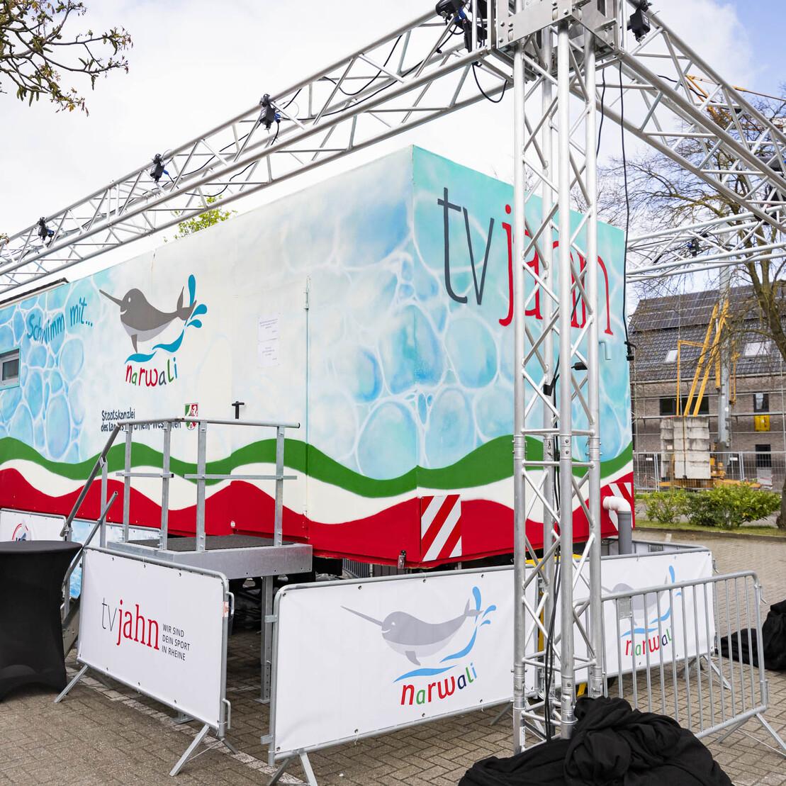 Mobiles Veranstaltungsgeb&auml;ude mit buntem Design und Absperrung auf einem Parkplatz, umgeben von B&auml;umen und Geb&auml;uden.